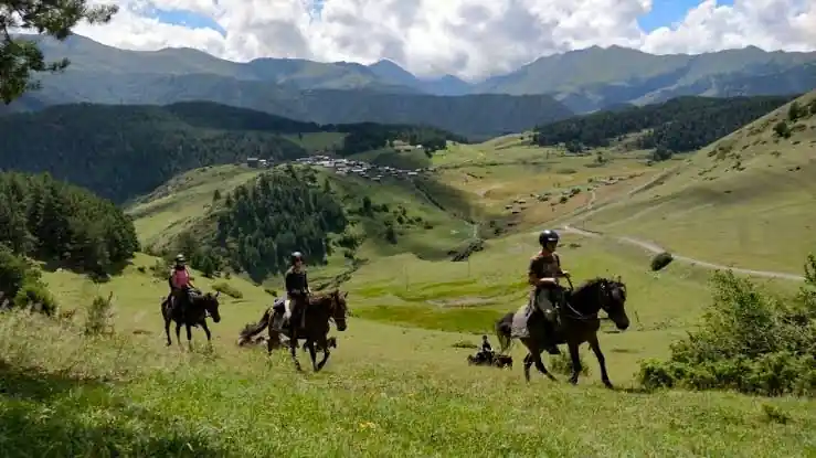 Drie ruiters op een groene bergweide in Toesjetië, met een torendorp in de verte